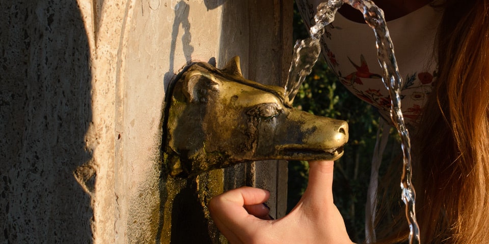 Ein Finger verschließt den Hauptauslauf eines Trinkbrunnens in der Villa Pamphilj, sodass das Wasser durch die obere Öffnung fließt