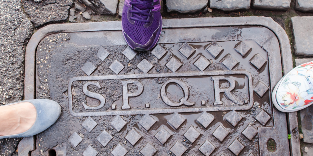Die Initialen SPQR auf einem metallenen Kanaldeckel in einer gepflasterten Straße in Rom